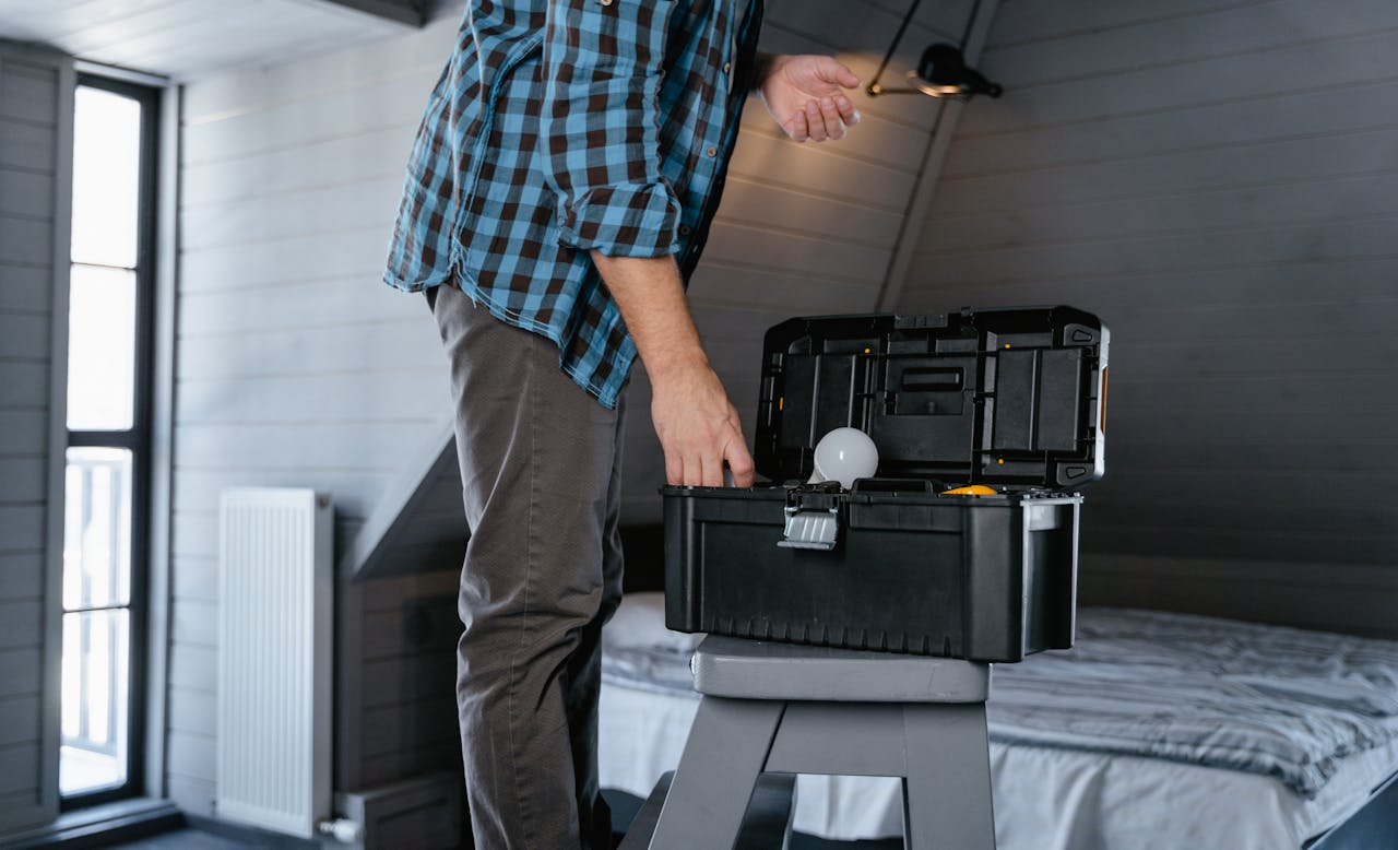 Strona główna Person in plaid shirt fixing a light bulb using a toolbox in a rustic attic interior.